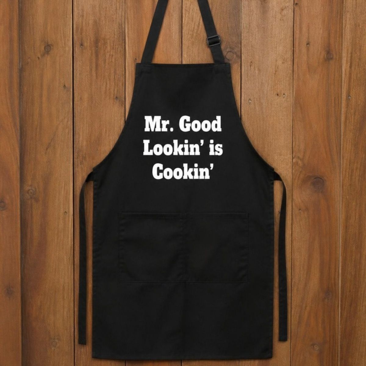 Black cotton apron with white text reading "Mr. Good Lookin' is Cookin'"
